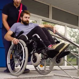 Mężczyzna na wózku inwalidzkim jest transportowany po schodach z pomocą drugiej osoby, widoczne wejście do budynku z niebieskimi drzwiami.