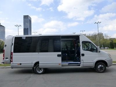 Biały bus z otwartymi drzwiami przesuwnymi na tle miejskiego krajobrazu z wieżowcami i drzewami.