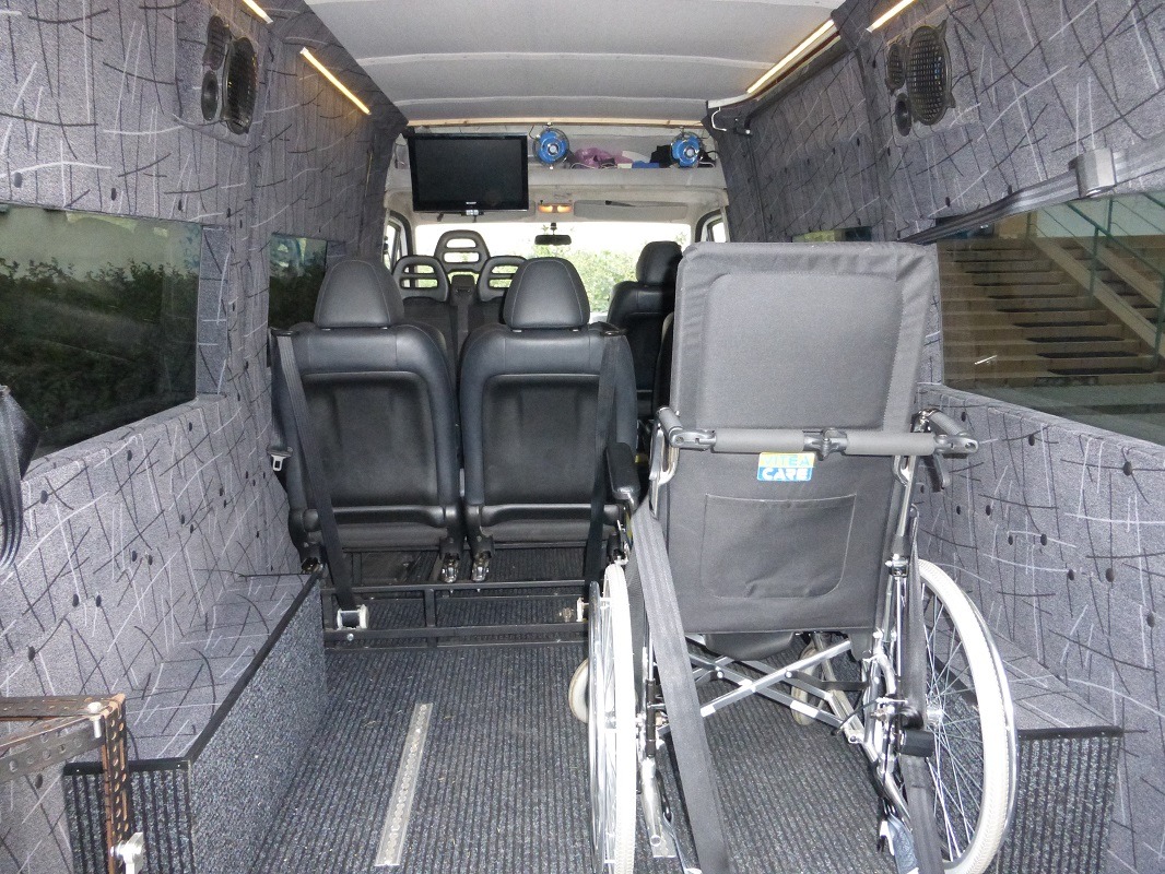 Wnętrze specjalistycznego busa przystosowanego do przewozu osób niepełnosprawnych, z zamontowanym wózkiem inwalidzkim, czarnymi skórzanymi fotelami, telewizorem i głośnikami w szarej tapicerce.