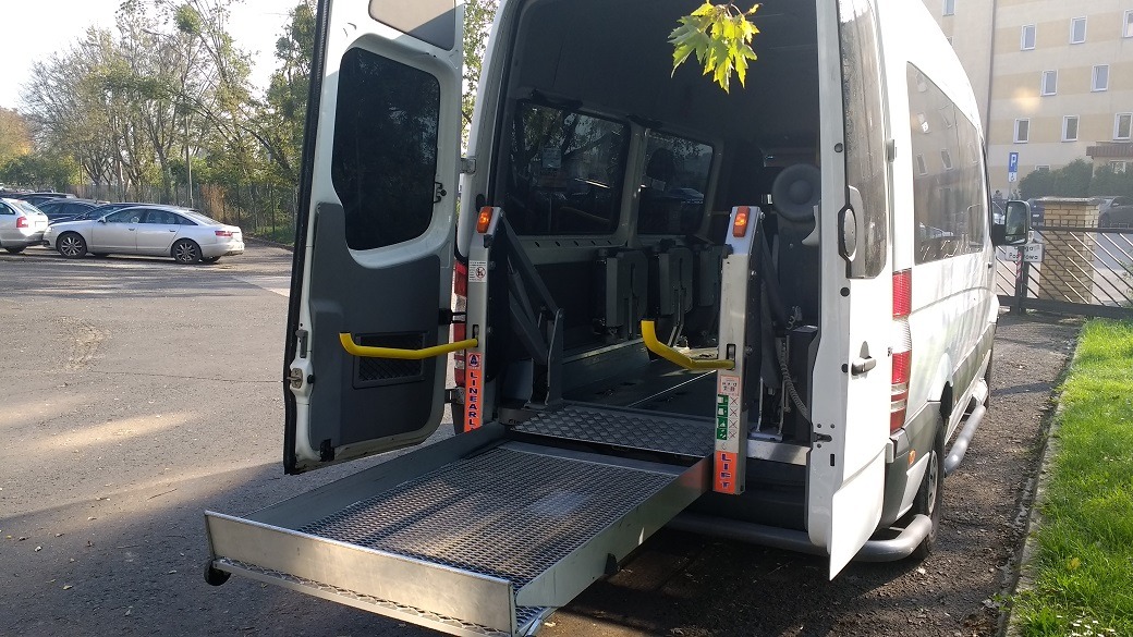 Biały bus z otwartymi drzwiami i rozłożoną platformą dla osób niepełnosprawnych na tle parkingu i budynku mieszkalnego.