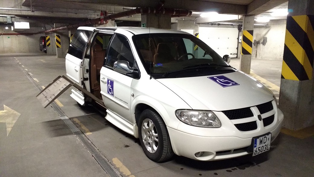 Biały van z rampą dla wózków inwalidzkich na parkingu podziemnym, otwarte drzwi boczne.