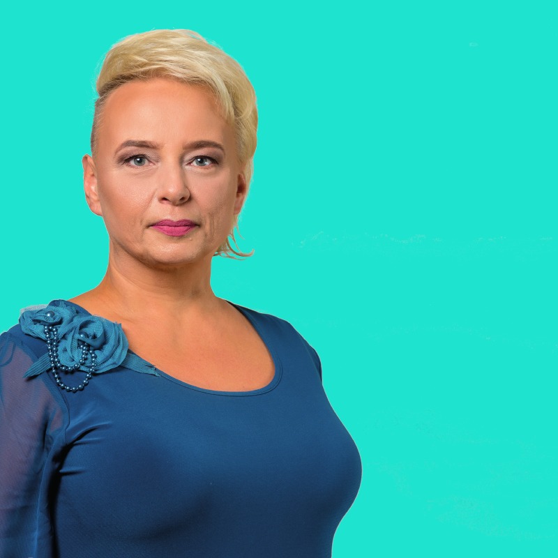 Portret kobiety z krótkimi blond włosami, ubrana w niebieską bluzkę z ozdobną różą na ramieniu, na jednolitym turkusowym tle.