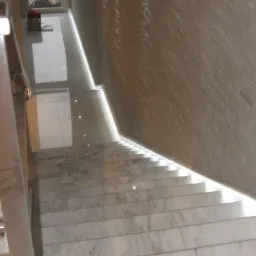 Marmurowe schody wewnętrzne z podświetleniem stopni, widok z góry na dół, ściana wykończona płytkami imitującymi kamień, widoczne prace wykończeniowe w tle za przeszklonymi drzwiami.