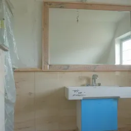 Łazienka w trakcie remontu: marmurowe płytki na ścianach, lustro w ramie z motywem greckim, biała umywalka z niebieską obudową zabezpieczoną folią, widoczne ślady po instalacji elektrycznej.