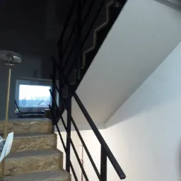 Wewnętrzne schody w trakcie budowy z surowymi stopniami z płyty OSB i czarną metalową balustradą, widok z dołu na okno z widokiem na domy.