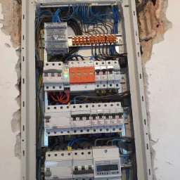 Otwarta skrzynka elektryczna z zainstalowanymi bezpiecznikami, przewodami i licznikiem energii elektrycznej, zamontowana na ścianie z widocznymi śladami po kuciu.