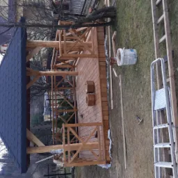 Wznoszona drewniana altana ogrodowa z ciemną dachówką bitumiczną, widoczne elementy konstrukcyjne, podłoga z desek, drabina i wiadro na trawniku.