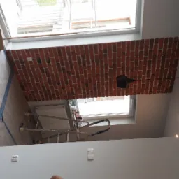 Częściowo wykończone wnętrze z dekoracyjną ścianą z cegły, zabezpieczone podłogi i drabina malarska wskazują na trwające prace wykończeniowe.