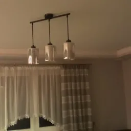 Trzy lampy wiszące z przezroczystymi kloszami na czarnej listwie, oświetlające wnętrze z oknem zasłoniętym firankami i zasłonami w odcieniach bieli i szarości.