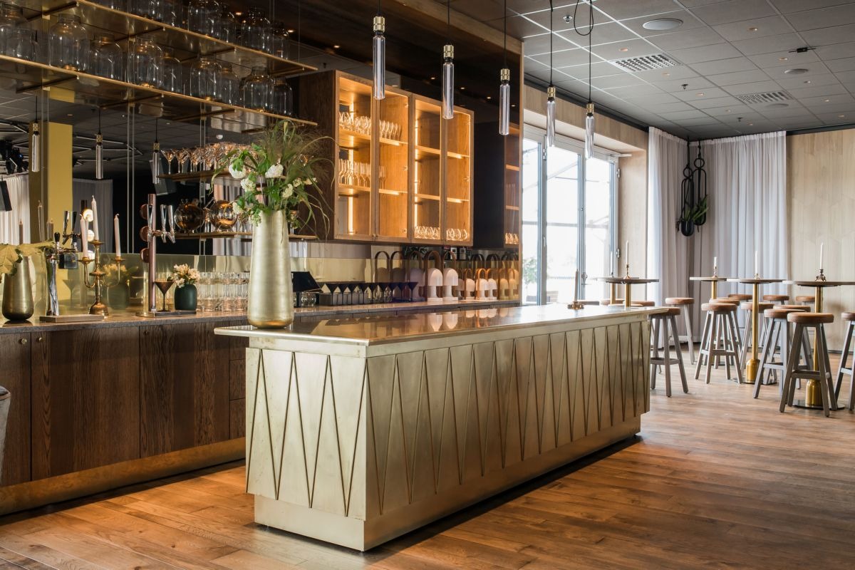 Nowoczesny bar w restauracji z drewnianą podłogą, złotym blatem z geometrycznym wzorem, szklanymi półkami pod sufitem i wiszącymi lampami.