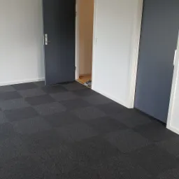 Pokój z szarą wykładziną dywanową w kratkę, białe ściany i dwie pary drzwi, jedne szare otwarte, drugie białe zamknięte.