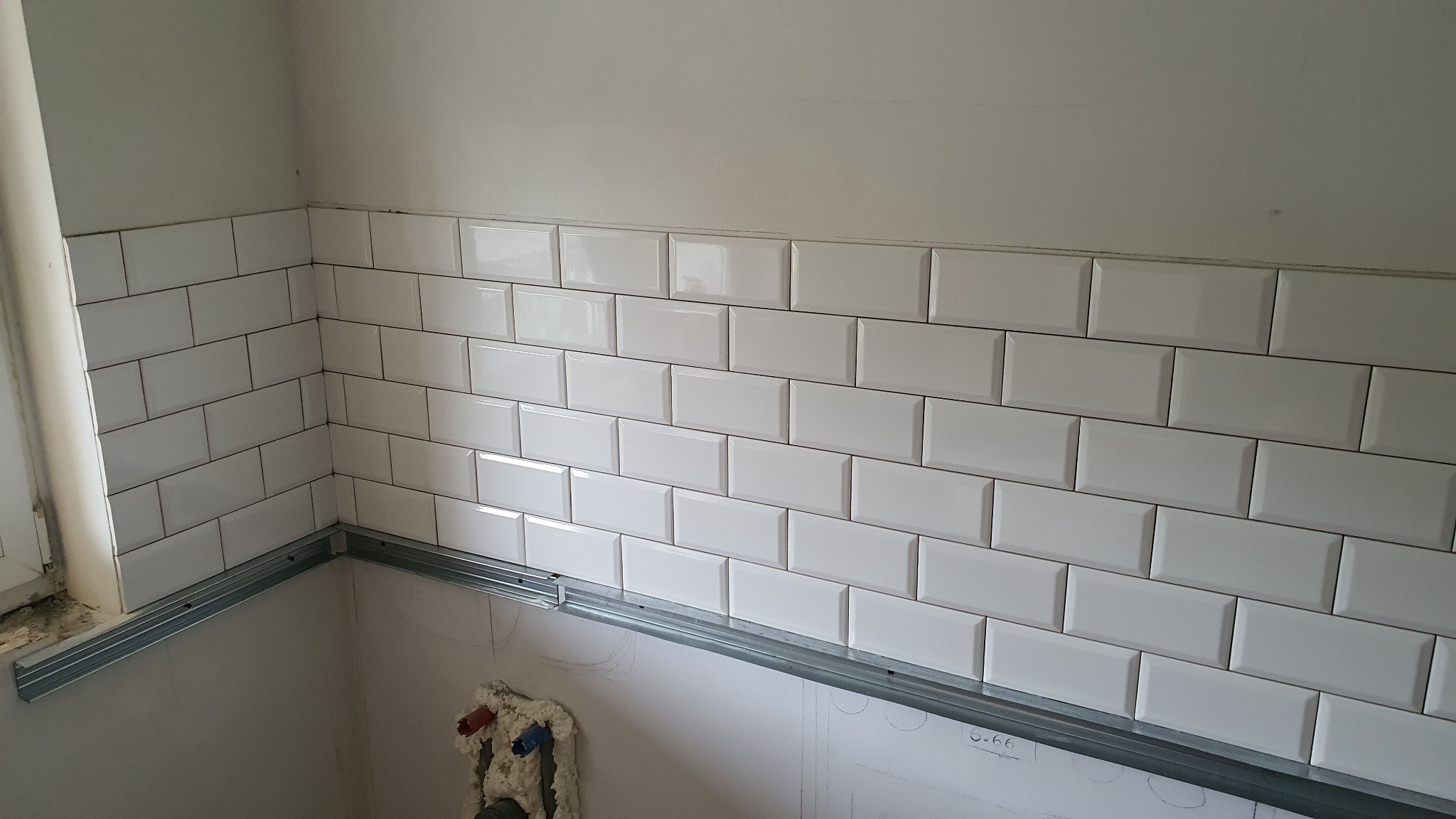 Ściana wyłożona białymi, błyszczącymi płytkami typu metro, z widocznymi metalowymi profilami montażowymi i fragmentem instalacji wodnej w pianie montażowej poniżej, w trakcie remontu łazienki.