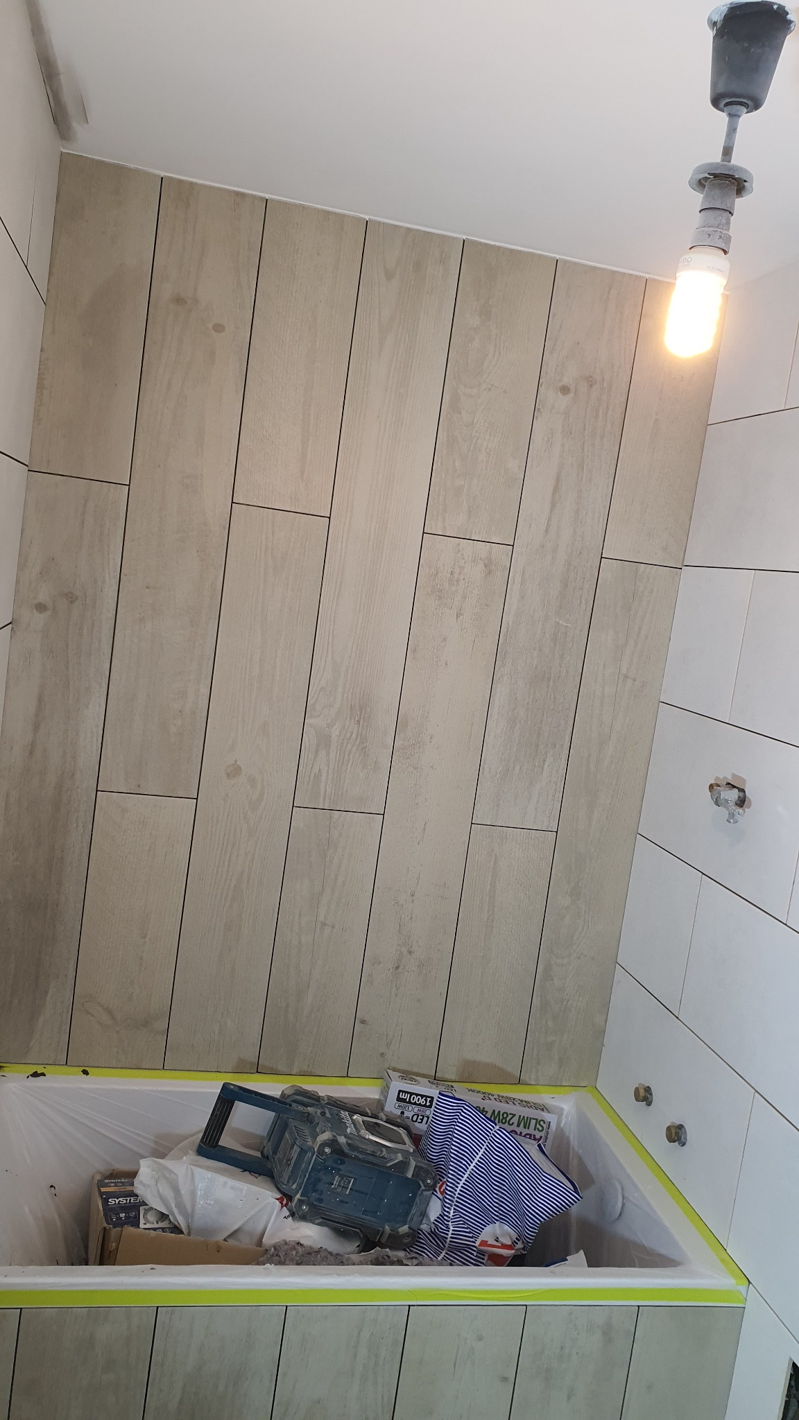 Ściana łazienki wyłożona płytkami imitującymi drewno, z widoczną wanną zabezpieczoną folią i taśmą, w której znajdują się narzędzia i materiały budowlane, pod wiszącą żarówką.