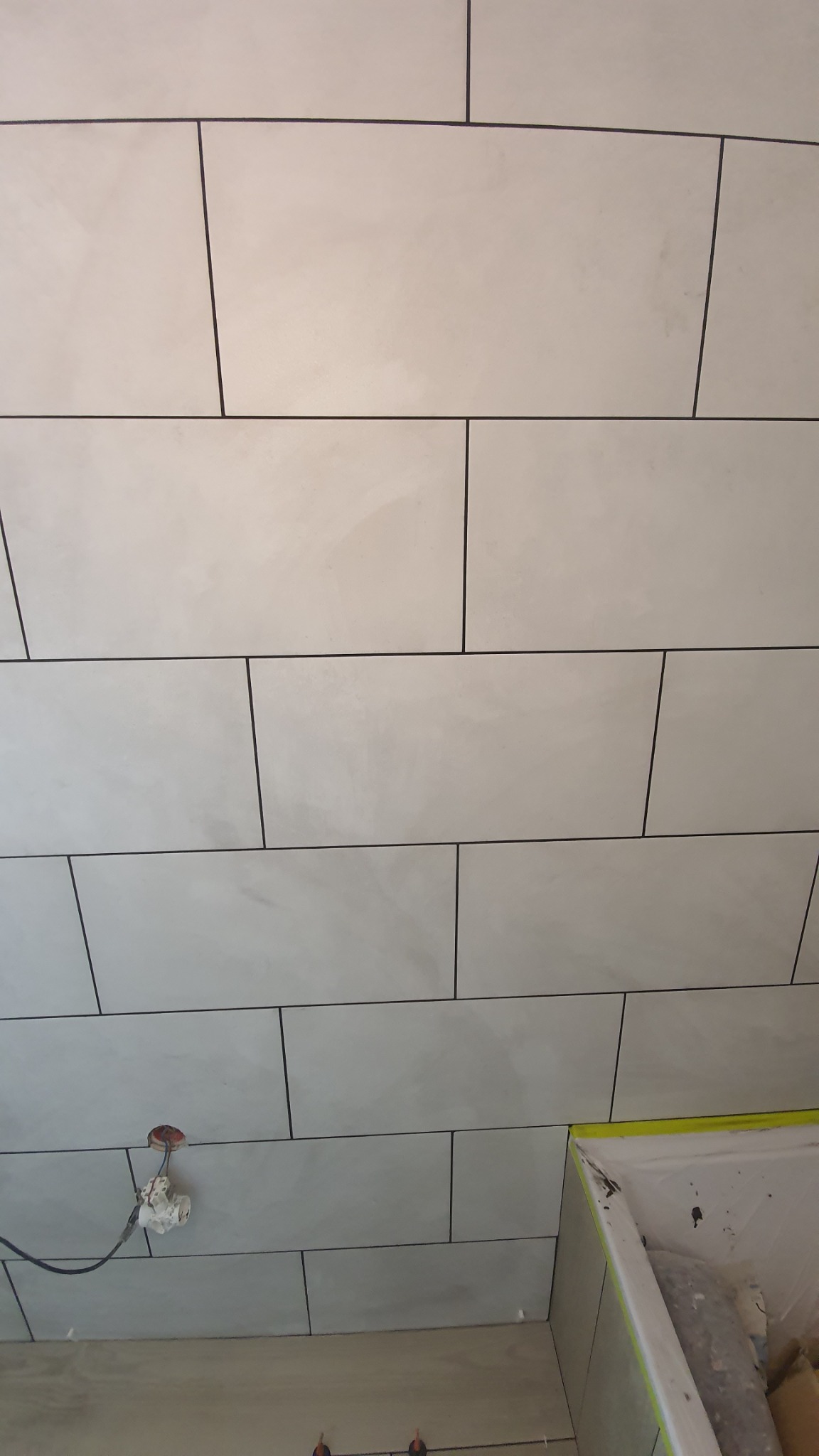 Ściana wyłożona podłużnymi, matowymi płytkami w odcieniach szarości z ciemnymi fugami, widoczna instalacja elektryczna i fragment zabezpieczonej folią wanny w trakcie prac.