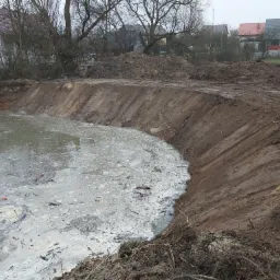 Nowo wykopany staw z mulistą wodą i strome, gliniaste brzegi. W tle widoczne zabudowania mieszkalne i nagie drzewa.