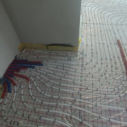 HYDROINSTAL Technika Grzewcza i Sanitarna - Rozłożona instalacja ogrzewania podłogowego z białych rurek na folii z siatką, widoczne czerwone i niebieskie rury przyłączeniowe. Ujęcie z góry, fragment ściany w tle.