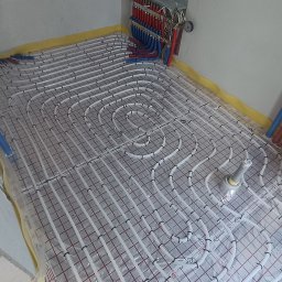 HYDROINSTAL Technika Grzewcza i Sanitarna - Białe rury ogrzewania podłogowego ułożone na folii z czerwoną kratką, widoczne kolektory z rurami czerwonymi i niebieskimi. Na ścianie notatki z instalacji.