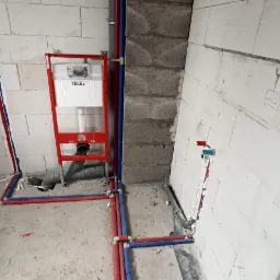 Surowy stan łazienki w trakcie remontu: widoczne instalacje wodno-kanalizacyjne z rurami w kolorach niebieskim i czerwonym, stelaż podtynkowy WC marki TECE na tle ściany z białego bloczka.