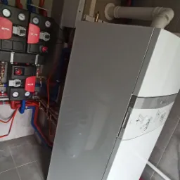 Nowoczesny, wiszący piec grzewczy z widocznymi, kolorowymi rurami instalacji CO i wodnej, oraz rozdzielaczami z manometrami, zamontowany na szarej, kafelkowanej ścianie.