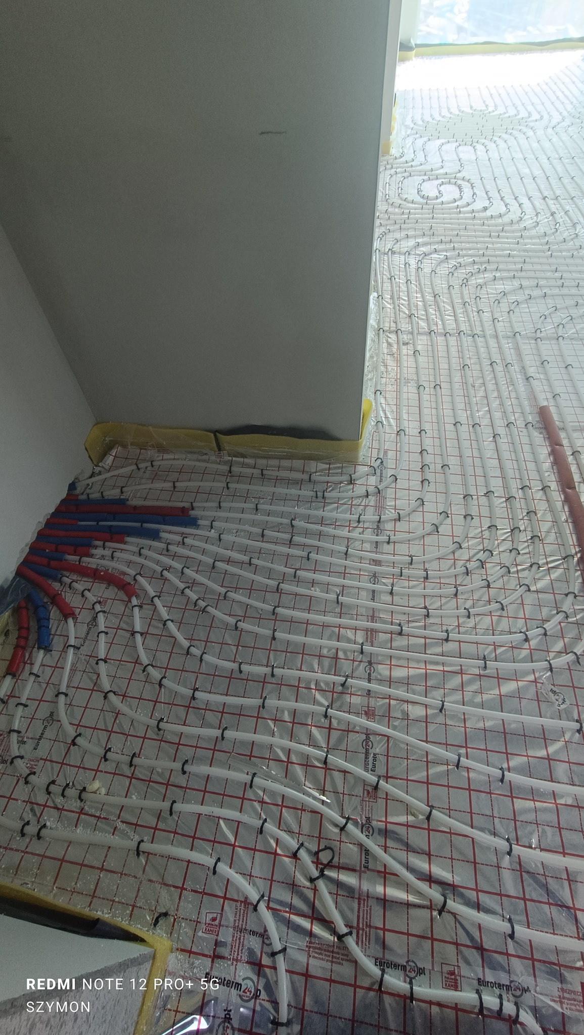 Rozłożona instalacja ogrzewania podłogowego z białych rurek na folii z siatką, widoczne czerwone i niebieskie rury przyłączeniowe. Ujęcie z góry, fragment ściany w tle.