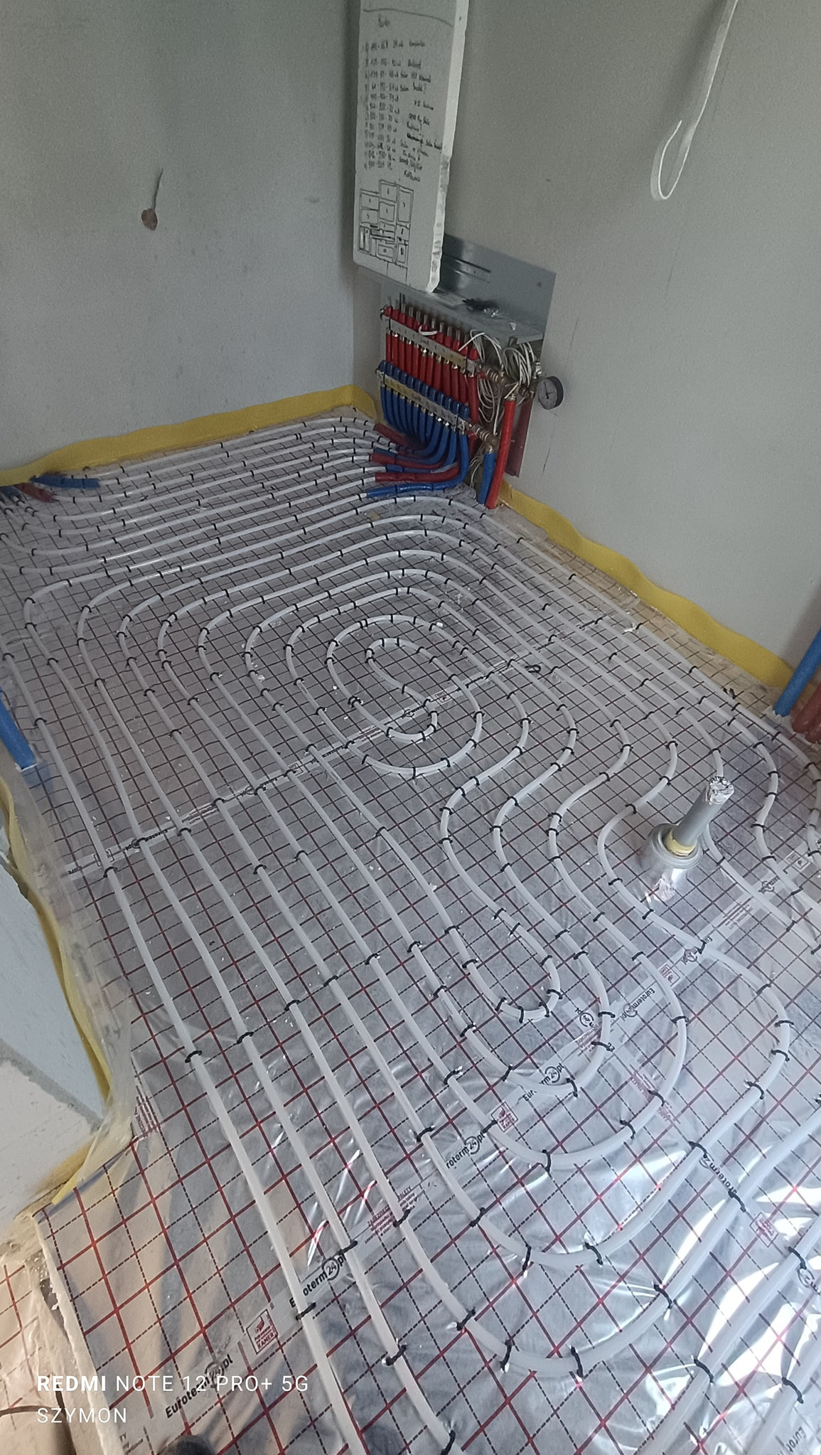 Białe rury ogrzewania podłogowego ułożone na folii z czerwoną kratką, widoczne kolektory z rurami czerwonymi i niebieskimi. Na ścianie notatki z instalacji.