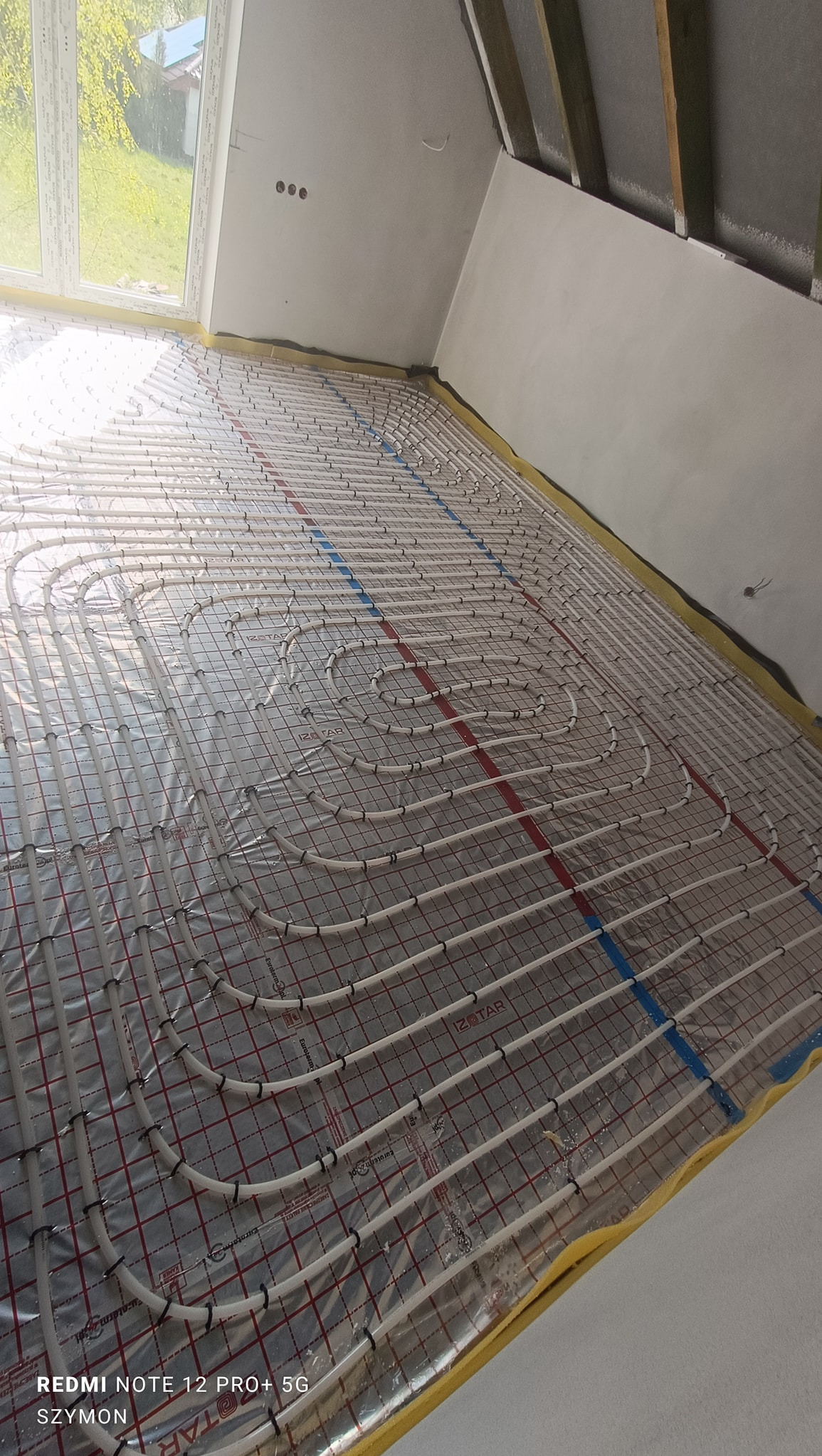 Ułożone rury ogrzewania podłogowego na folii izolacyjnej z siatką, widok z góry, przed wylaniem posadzki. Żółta taśma brzegowa wzdłuż ścian.