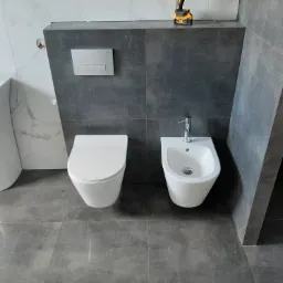 Nowoczesna łazienka z białymi sanitariatami (umywalka na postumencie, toaleta, bidet) i szarą glazurą, widoczny wiertarko-wkrętak na górnej półce.