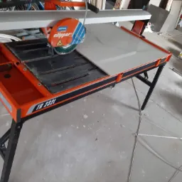 Elektryczna przecinarka do płytek Norton Clipper TR 232I z płytką ceramiczną gotową do cięcia, w tle wiadro z zaprawą i inne akcesoria.