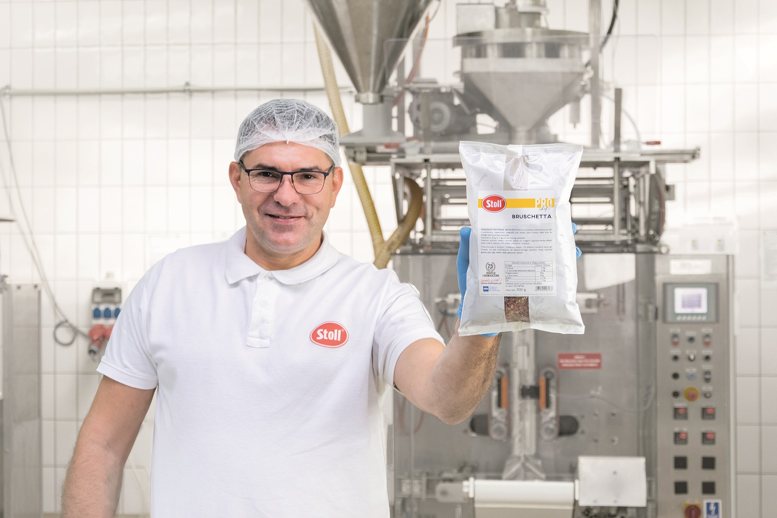 Pracownik w czepku i rękawicach prezentuje opakowanie mieszanki przypraw do bruschetty marki Stoll na tle linii produkcyjnej.