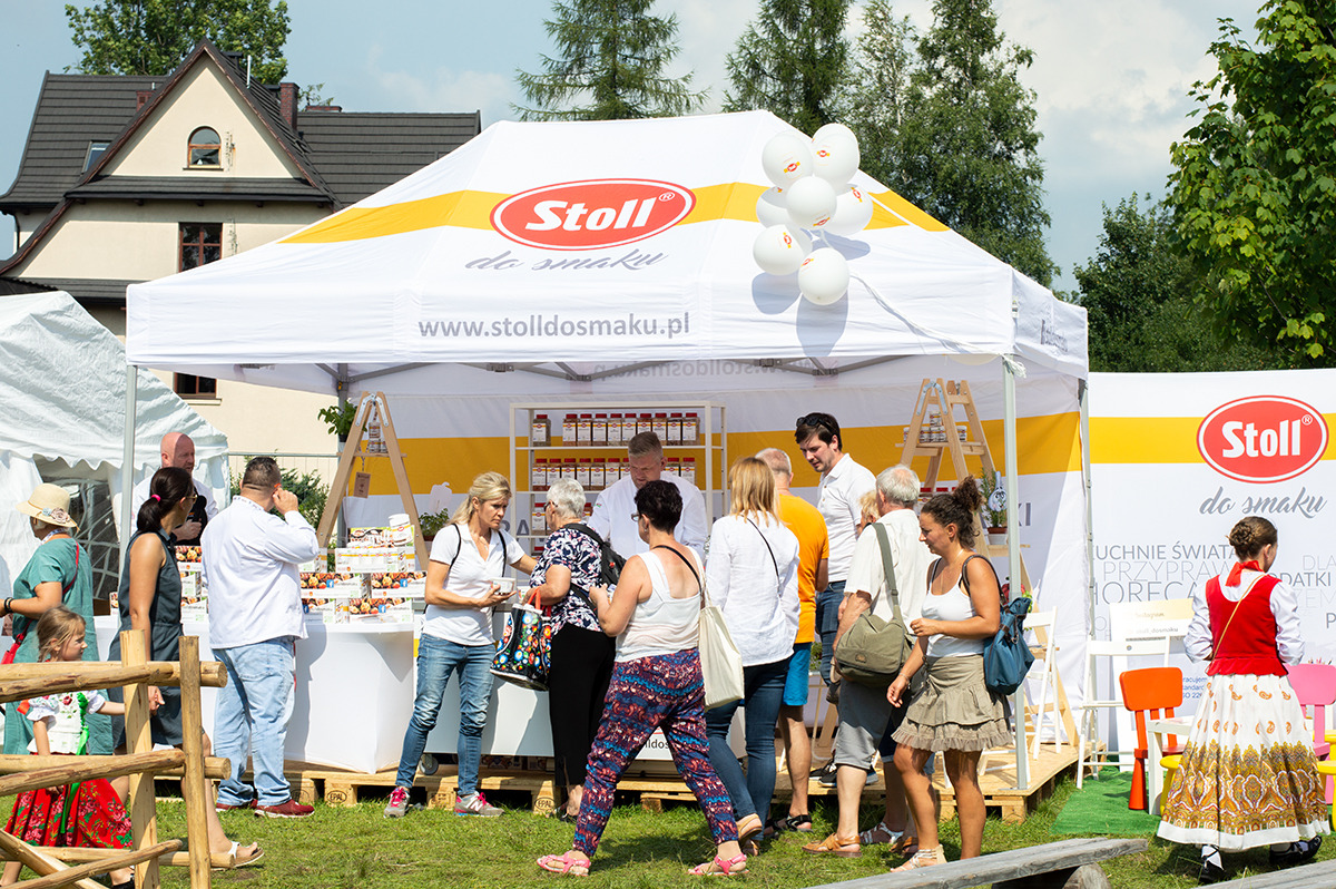 Stoisko z produktami spożywczymi marki Stoll na plenerowym wydarzeniu, wokół którego gromadzą się klienci. Widoczne białe namioty z logo, balony i osoby w różnym wieku.