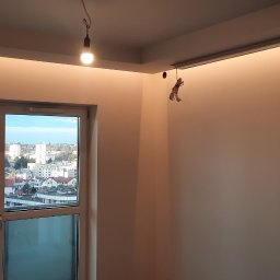 Pracuś Remonty I Wykonczenia - Pomalowane na biało wnętrze pokoju z dwoma oknami z widokiem na miasto, sufit z ukrytym oświetleniem led, wisząca żarówka na kablu.