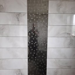 Pracuś Remonty I Wykonczenia - Ściana wyłożona płytkami imitującymi marmur z pionowym pasem mozaiki w odcieniach szarości, z widocznym odbiciem fotografa.