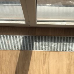 Pracuś Remonty I Wykonczenia - Szczelina wentylacyjna w podłodze z drewnianych paneli, zabezpieczona folią malarską podczas prac remontowych, widok z góry, okno w tle.