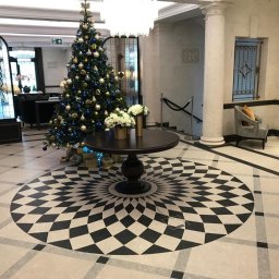 Pracuś Remonty I Wykonczenia - Eleganckie lobby hotelowe z dekoracyjną posadzką w geometryczny wzór, okrągłym stolikiem z kwiatami i świąteczną choinką z niebieskimi i złotymi ozdobami.