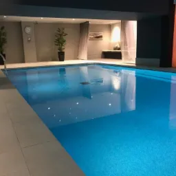 Kryty basen z błękitną wodą, wykończony jasnymi płytkami, z roślinami w donicach i zegarem na ścianie w tle.