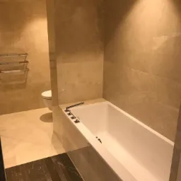 Wykończona łazienka z beżowymi marmurowymi płytkami na ścianach i podłodze, biała wanna z chromowaną armaturą, widoczny fragment toalety i ciemnobrązowa marmurowa podłoga przy wejściu.