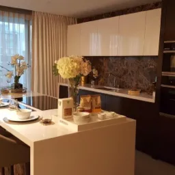 Nowoczesna kuchnia z wyspą, barkiem i krzesłami barowymi, marmurowy panel nad blatem, białe szafki górne i ciemne dolne, zastawa stołowa i dekoracje na blacie, widok na okno z zasłonami.