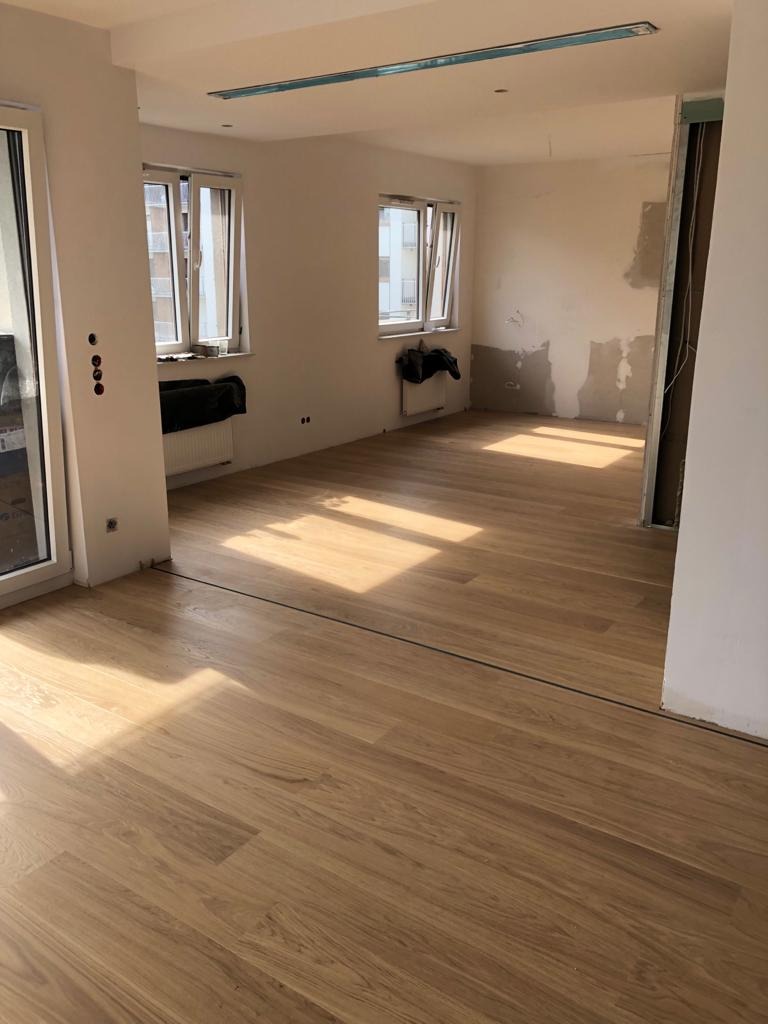 Jasna drewniana podłoga w pokoju po remoncie, widoczne ślady gładzi na ścianach, okna z grzejnikami, listwa sufitowa z oświetleniem.