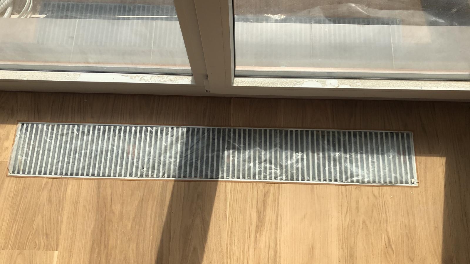 Szczelina wentylacyjna w podłodze z drewnianych paneli, zabezpieczona folią malarską podczas prac remontowych, widok z góry, okno w tle.