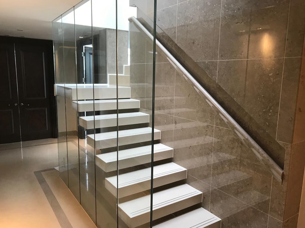 Nowoczesne schody z białymi stopniami i szklaną balustradą, obłożone płytkami imitującymi kamień, z widocznym fragmentem ciemnych drzwi w tle.