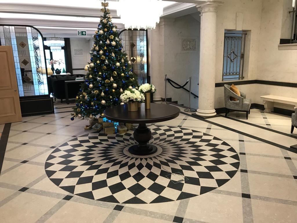 Eleganckie lobby hotelowe z dekoracyjną posadzką w geometryczny wzór, okrągłym stolikiem z kwiatami i świąteczną choinką z niebieskimi i złotymi ozdobami.