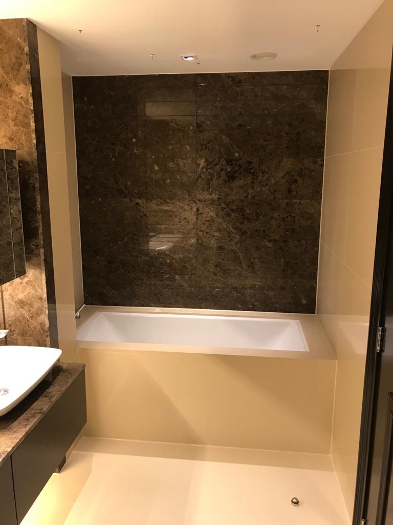 Nowoczesna łazienka z wanną obudowaną beżowymi płytkami, ściana nad wanną wyłożona ciemnym marmurem, umywalka nablatowa z szafką z blatem imitującym marmur.