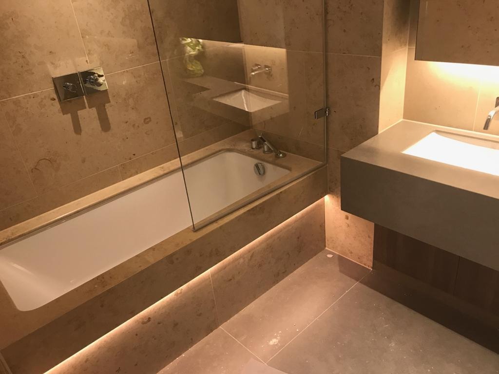 Nowoczesna łazienka z beżowymi płytkami imitującymi marmur, podświetlana wanna z parawanem i prostokątna umywalka z szafką w zabudowie.