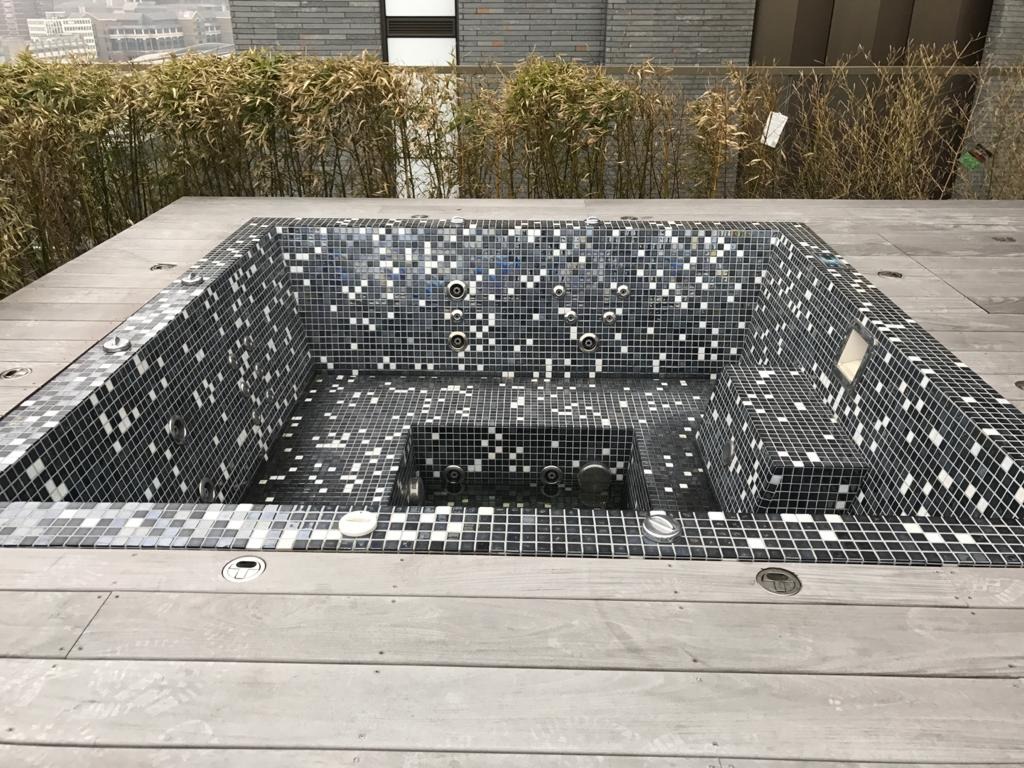 Zewnętrzna wanna z hydromasażem wyłożona czarno-białą mozaiką, otoczona drewnianym tarasem i bambusową roślinnością w tle.