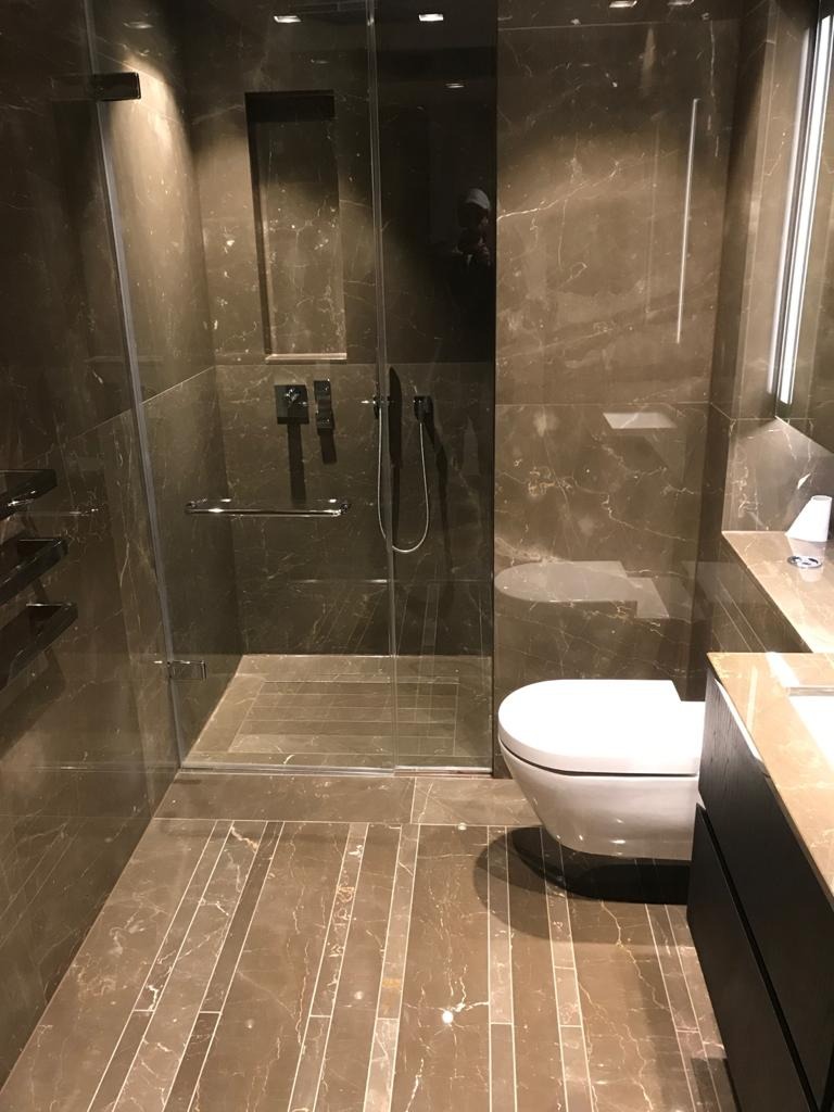 Nowoczesna łazienka z kabiną prysznicową typu walk-in, wykończona płytkami imitującymi marmur w odcieniach brązu, z subtelnym oświetleniem LED i toaletą podwieszaną.