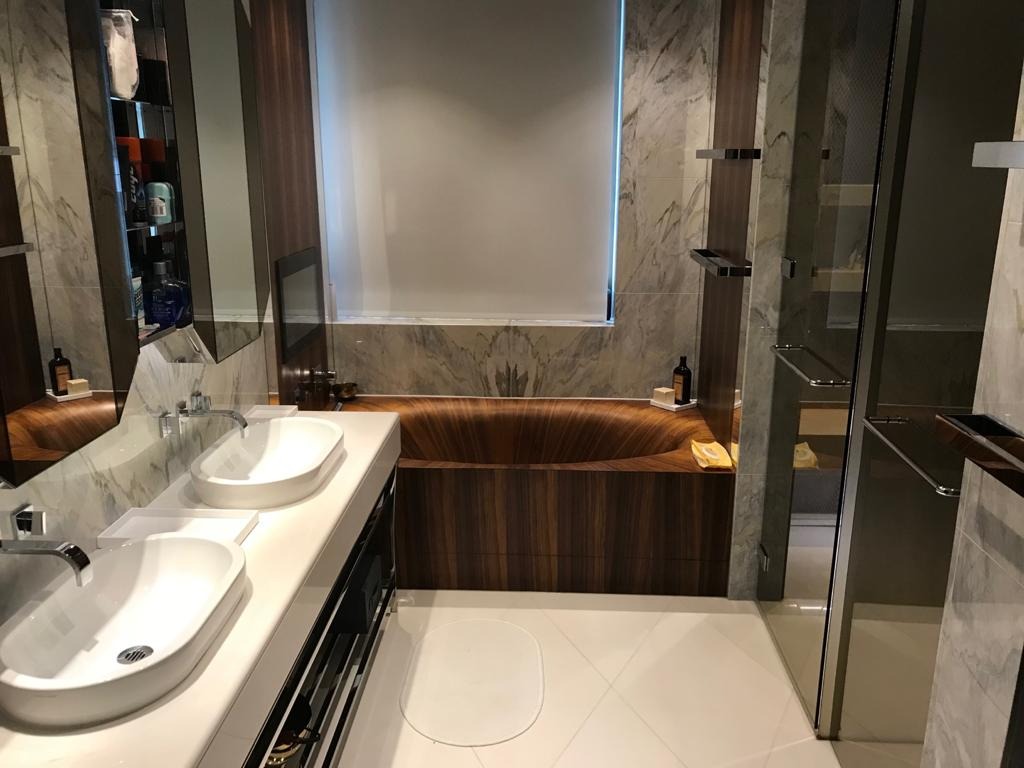 Nowoczesna łazienka z drewnianą wanną w zabudowie, marmurowymi ścianami i podwójną umywalką na blacie, widoczna kabina prysznicowa i lustro z półkami.