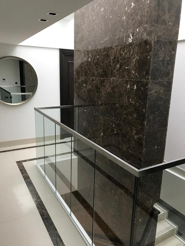 Wykończenie klatki schodowej z przeszkloną balustradą i kolumną obłożoną płytami imitującymi ciemny marmur z jasnymi żyłami, okrągłe lustro na ścianie.