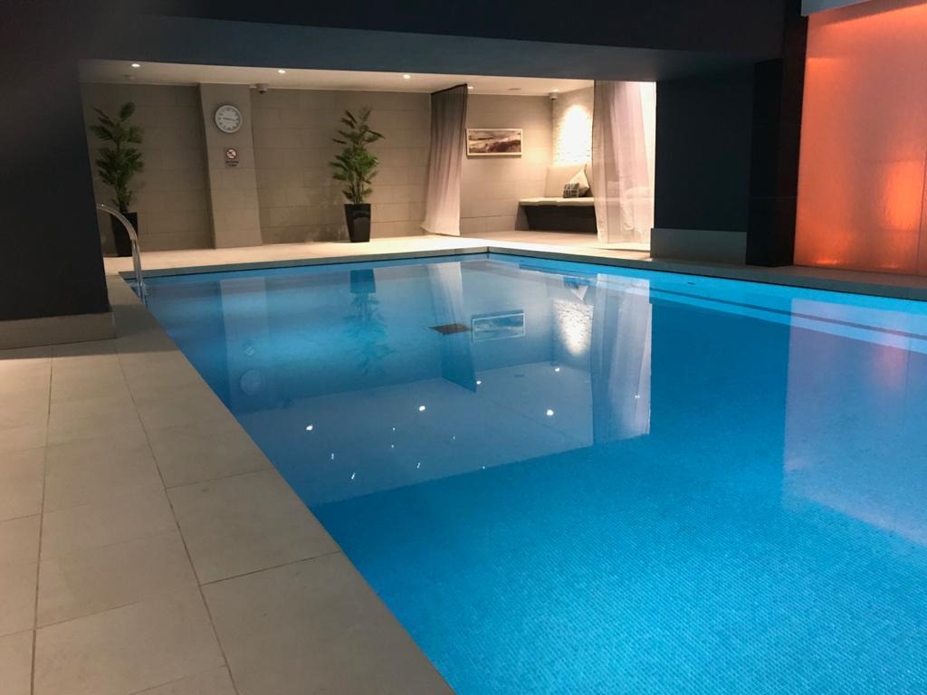 Kryty basen z błękitną wodą, wykończony jasnymi płytkami, z roślinami w donicach i zegarem na ścianie w tle.