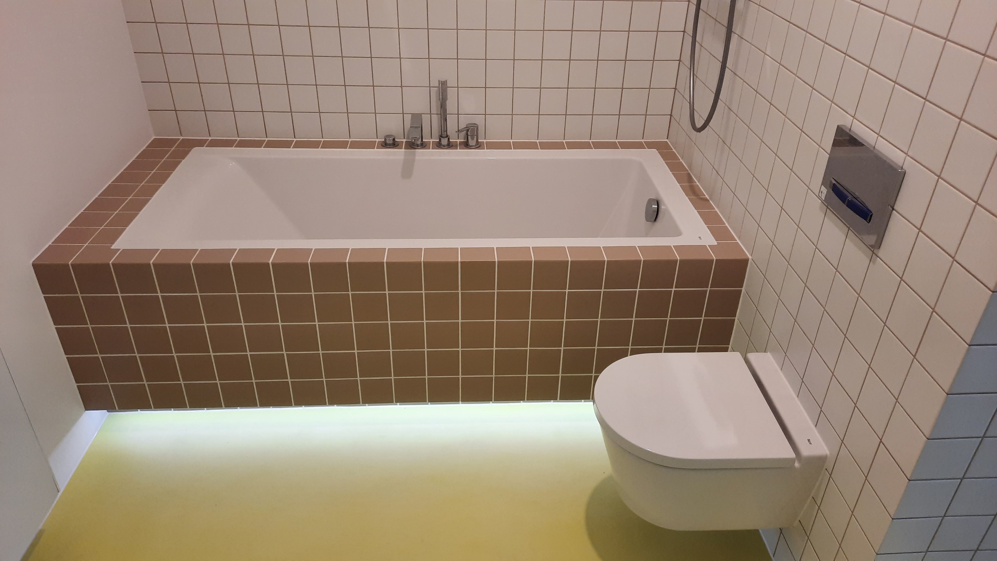 Biała wanna obudowana brązowymi płytkami, wisząca toaleta z podświetleniem LED pod wanną na żółtej podłodze, białe kafelki na ścianach.