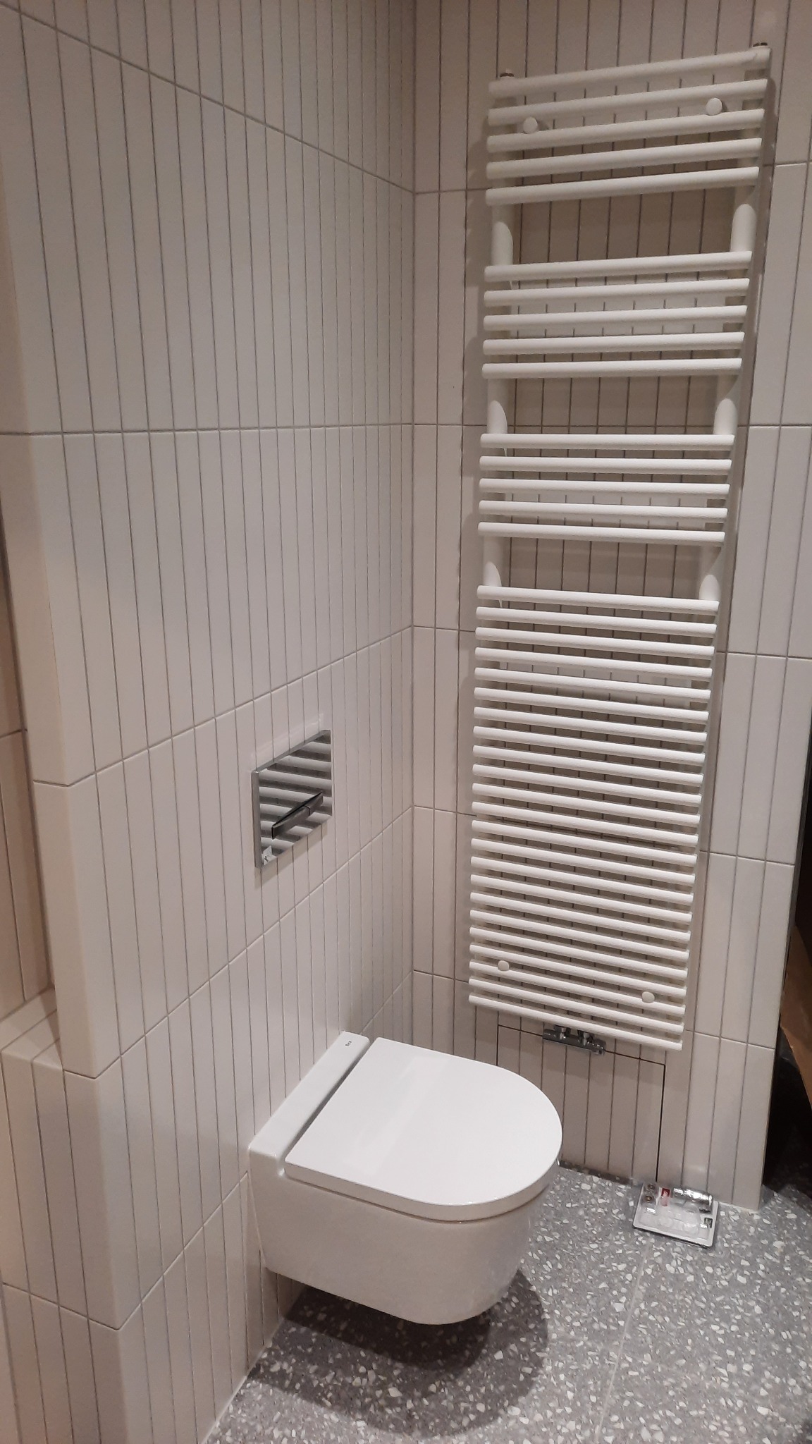 Nowoczesna, podwieszana toaleta z chromowanym przyciskiem spłukującym i białym grzejnikiem drabinkowym na ścianie wyłożonej pionowymi, kremowymi płytkami, szara posadzka z terazzo.